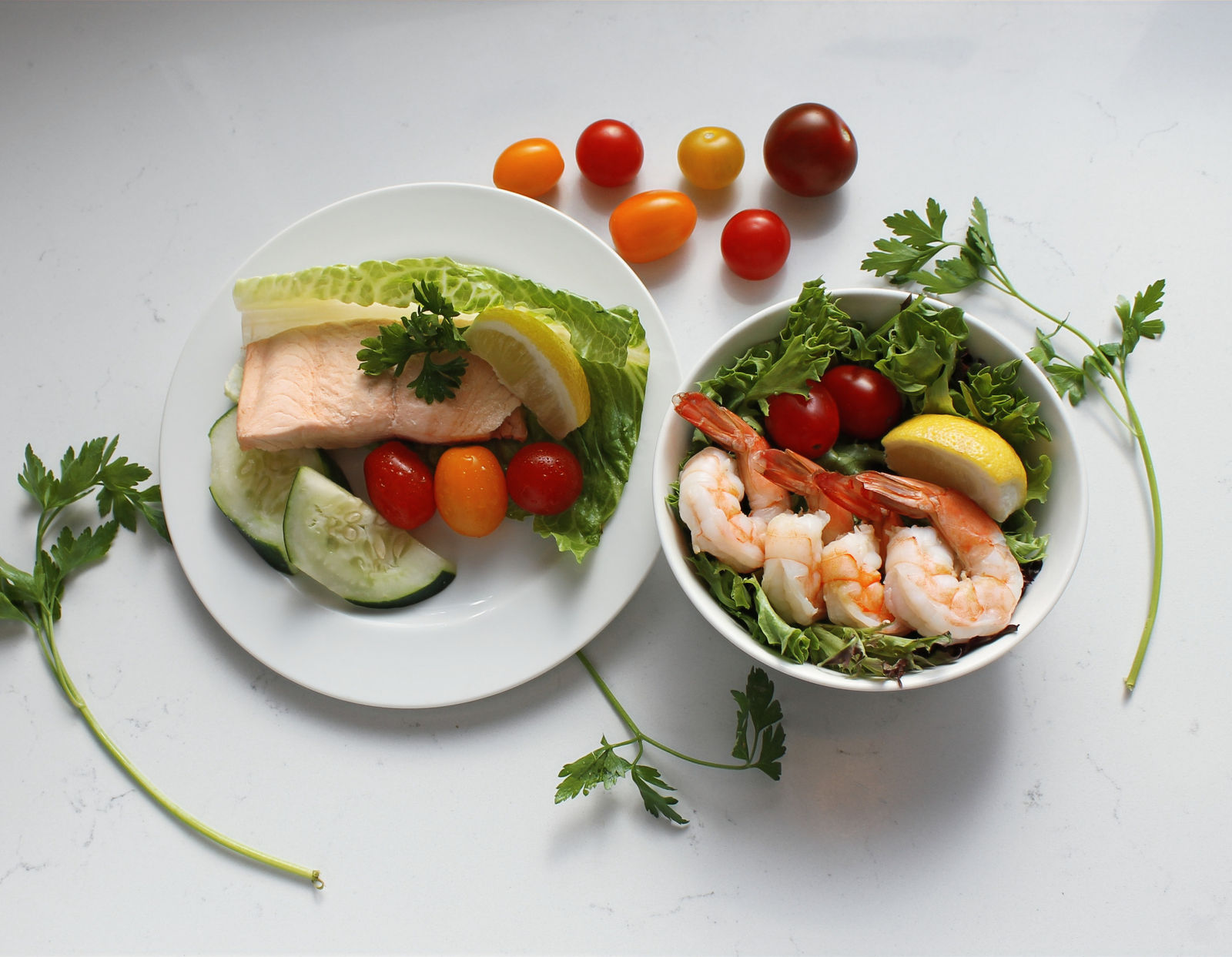 A salmon salad with lemon and a shrimp and tomato salad on a counter scattered with cherry tomatoes and parsley